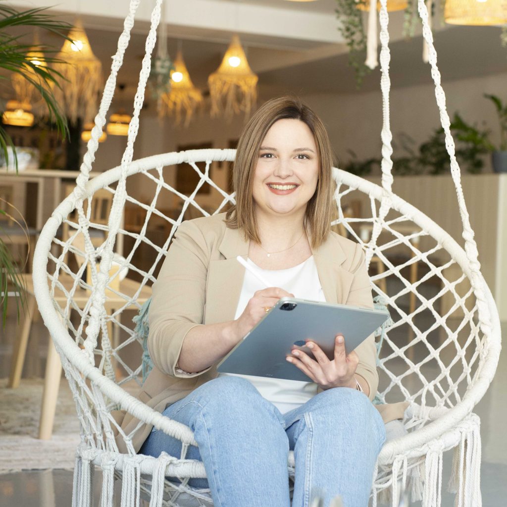 Sylvia in einem Hängestuhl mit iPad und Apple Pen in den Händen und Blick auf die betrachtende Person.